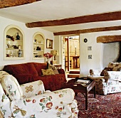 Niedriges Wohnzimmer mit weiss verputzter Holzbalkendecke und rotem Sofa vor eingebauten Wandregalen neben Sesseln in floralem Muster