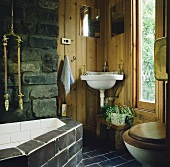 Ein schwarzer Fliesenboden umgibt ein rustikales Badezimmer mit einem Eck-Waschbecken, das auf einer holzgetäfelten Wand neben einer Steinmauer befestigt wurde