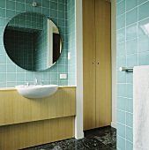 In einem türkis gefliesten Bad hängt ein kreisförmiger Spiegel über einem Waschbecken, das in einer holzgetäfelten Einheit eingelassen ist