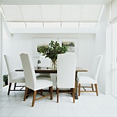 Weisses Esszimmer mit altem, quadratischem Holztisch und gepolsterten Stühlen unter Glasdach mit Stoffjalousie