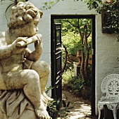 Flötespielender Steinengel in kleinem Innenhof und antikem Metallstuhl neben Blick in den Garten durch offene Tür