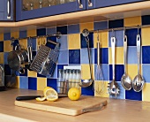 Küchenutensilien an einer Stange mit Fleischerhaken aufgehängt vor gelb und blau gefliester Rückwand