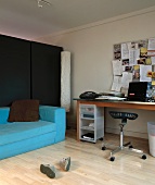 Modernes Wohnzimmer mit pastellblauem Matratzensofa vor schwarzer Trennwand und Schreibtisch mit vollgehängter Pinnwand