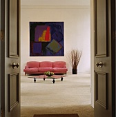Ein Couchtisch aus Glas und ein pinkfarbenes Sofa unter einem abstrakten Bild