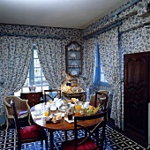 Esszimmer mit blau-weiss gemusterten Fenstervorhängen, den dazu passenden Tapeten und einem gedeckten Frühstückstisch