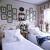 Zwei Einzelbetten mit gepolsterten Kopfteilen mit Blumenmotiv und weisser Bettwäsche, darüber eine Sammlung gerahmter Pflanzendrucke