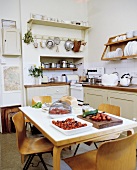 Essensvorbereitung auf Küchentisch mit Holzstühlen im Retrostil vor ländlicher Küchenzeile mit hellgrauen Fronten