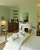weiße Kissen und Decke auf Messing-Bett und antiker Kommode neben Kamin in einem lindgrünen Schlafzimmer