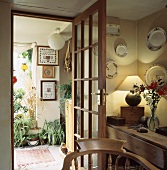 Zimmerecke mit Wandtellern im Kunstlicht der Tischlampe neben offener Glastür mit Blick auf Pflanzen im Vorraum