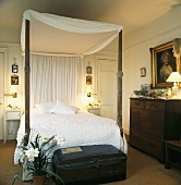Himmelbett mit weißem Baldachin und dunklem Holzgestell mit alter Truhe am Bettende