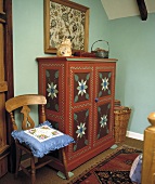 Bemalter Schrank im bäuerlichen Stil und Stuhl mit Kissen vor hellblauer Wand