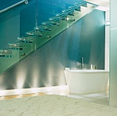 A free standing bathtub underneath a flight of glass stairs