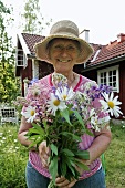 Ältere Frau mit Blumenstrauss im Garten vor Haus