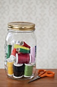 Assorted spools of thread in a preserving jar, sewing scissors next to it
