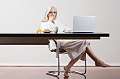 Frau mittleren Alters im Bademantel sitzt mit Frühstück und Laptop an Arbeitstisch