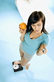 Young woman standing on scales with chocolate bar & orange