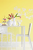 Two vases of tulips and cake stand of confectionary on white table in front of yellow wall
