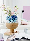 A glass container filled with blue Easter eggs, sprigs of blossom and buttercups