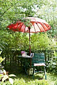 Seating area in summery garden with garden chair, garden table & red Thai-style parasol