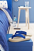 Blue-and-white flip-flops with crocheted straps in front of bed with blue bedclothes
