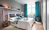 A bedroom with light-grey walls and blue curtains with a long bookshelf over a double bed