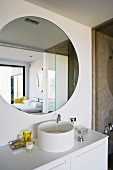 A white designer wash stand with bathing utensils and a round wall mirror