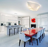 Open-plan dining room with red painted dining table and white upholstered chairs in front of kitchen counter and bar stools