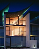Modernes Gebäude mit abendlich beleuchteter Glasfassade und segelförmig aufgebogenem Dach mit grünem Lichteffekt