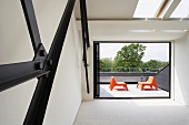 Blick von leerem Innenraum mit Metalltragkonstruktion durch offene Fensterfront auf Dachterrasse mit Sesselpaar aus orangefarbenem Kunststoff