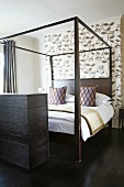 Double bed with four-poster frame and scatter cushions in front of patterned wallpaper in modern bedroom