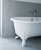 Freistehende Badewanne mit Klauenfüssen und moderner Armatur auf grauen, matten Fliesen vor grauer Wandverkleidung