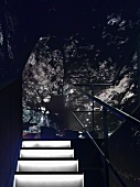 Treppenaufgang mit weiss beleuchteten Stufen und einfachem Metallgeländer mit undeutlichem Blick auf Essplatz unter grau-schwarzen Strukturen