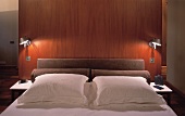 Modern double bed with lit wall lamps and free-standing wooden rear wall