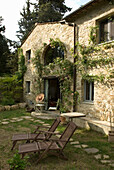 Steinhaus mit Rosenranken und Holzliegestühlen im Garten