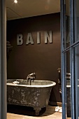 Blick durch offene Glastür mit Metallrahmen auf Vintage Badewanne mit Füssen vor dunkelbraun getönter Wand und aufgesetzten Buchstaben