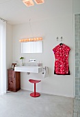 Schlichtes Bad mit modernem Waschbecken und rotem Hocker neben Kleid an Wandhaken gehängt