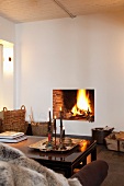 Sofa und Tisch mit Kerzen vor schlichter Kaminöffnung mit loderndem Feuer in glatter weisser Wand