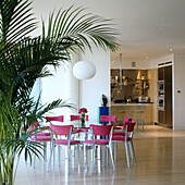 Dining area with pink chairs and palm tree, open kitchen in the background