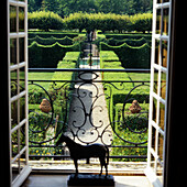 View through open balcony doors to a formal garden with boxwood hedges