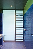 Frosted glass wall behind floor-level shower in bathroom with blue mosaic tiles under green glass strip