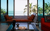 Wooden recamier with loose cushions in front of large windows with view of wooden terrace and palms behind infinity pool