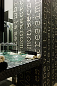 Running water from a wall mount faucet into a glass bowl on a designer vanity with a tile mosaic made of a dark background with white lettering