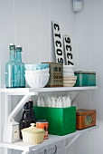Crockery, swing-top bottles, candles, number plates and tins on white, wall-mounted shelves