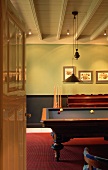 View through open door of wooden billiard table in simple room with white wooden ceiling and red floor