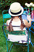 Girl with pigtails and straw hat sits on vintage garden chair in front of flower decoration