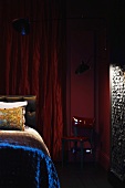 Corner of red-painted bedroom in semi-darkness - bed with shiny bedspread in front of red curtain on wall