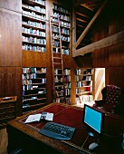 Ceiling-height fitted shelving reaching into open roof structure and antique desk with monitor and keyboard