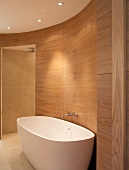 Free-standing bathtub against wood-panelled wall in oval, modern bathroom