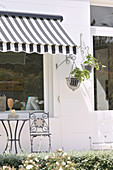 Small table and chair on terrace below black and white striped awning on whitewashed house