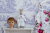 Doll's head and rabbit soft toy on shelf against Toile de Jouy wallpaper